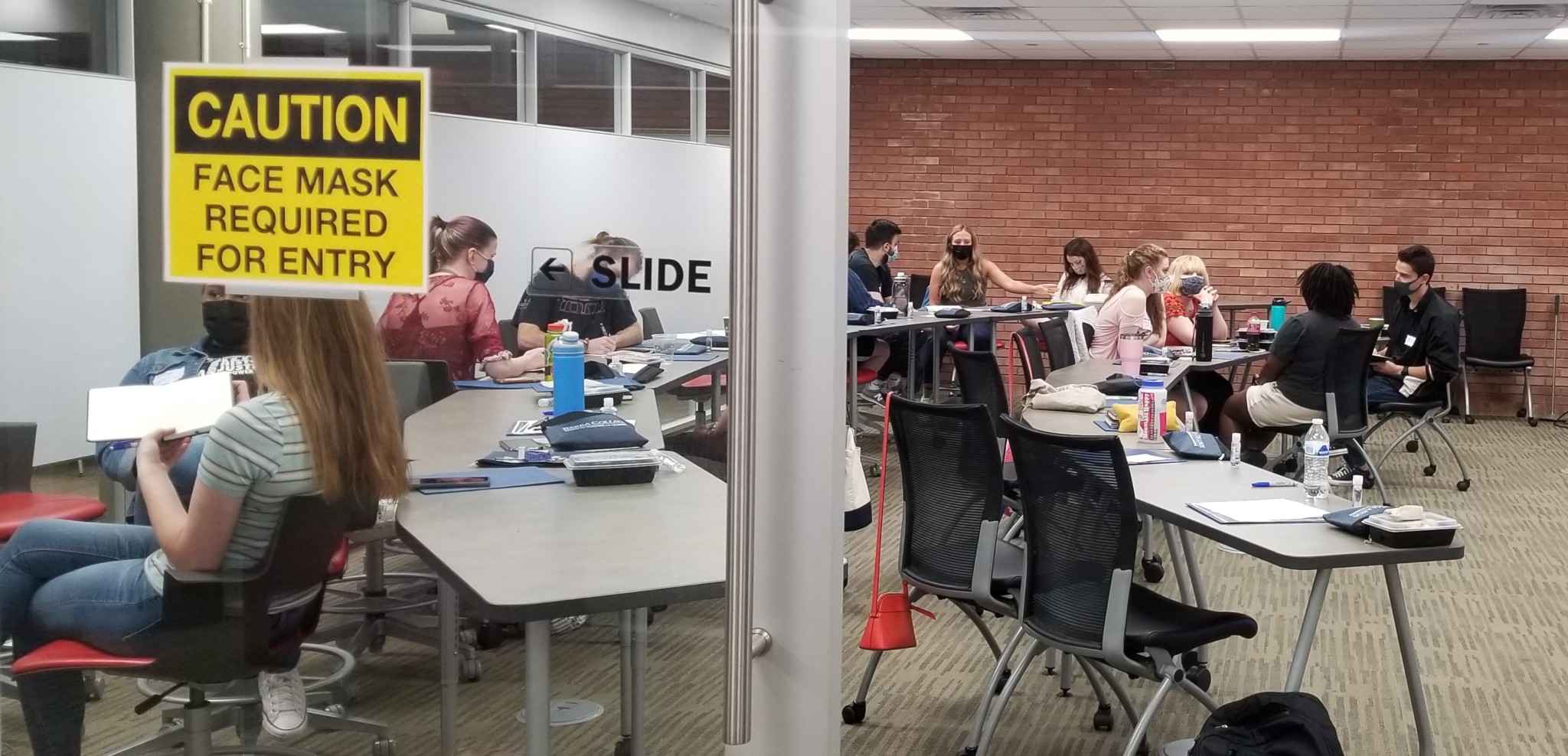 20 college students are in a classroom, divided into groups. They are working together. Some are writing. Some are talking. Some are listening.